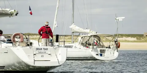 Un homme en gilet rouge tient la barre d'un voilier, le pont est bien mouillé. Un autre voilier suit, naviguant sur une mer calme.