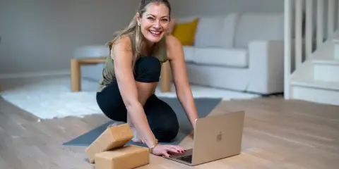 Glad kvinna förbereder sig för hatha yoga hemma, med yogablock och laptop redo.