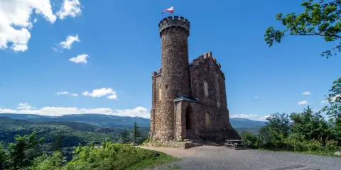 Kamienny zamek księcia Henryka na trasie widokowej, z polską flagą na wieży, otoczony zielonymi wzgórzami.
