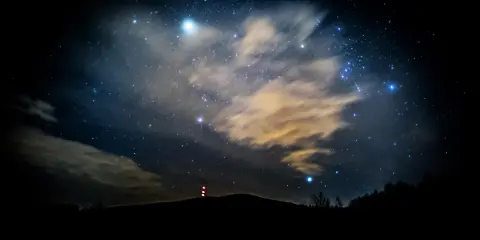 Nocne niebo pełne gwiazd, chmur i odległych świateł. W oddali widać zarys wzgórza z wieżą. To właśnie takie widoki towarzyszą nam, gdy zastanawiamy się, co to są Perseidy.