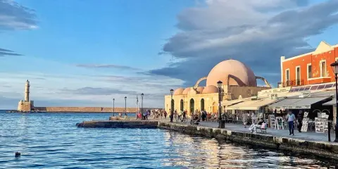Il porto di Chania con il suo faro iconico e la moschea ottomana, bagnato da un mare blu intenso sotto un cielo nuvoloso.