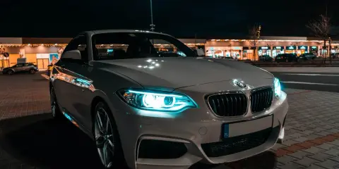 Białe BMW z włączonymi światłami adaptacyjnymi błyszczy nocą przed sklepami.