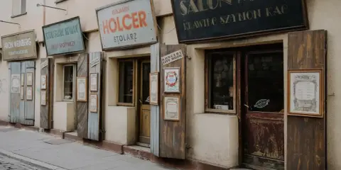 Ulica w dzielnicy żydowskiej Krakowa z zabytkowymi szyldami sklepów: "Salon Mód", "Benjamin Holcer Stolaz", "Stanisław Nowak Sklep Spożywczy".