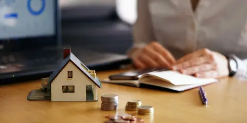 Maqueta de casa, monedas y portátil con gráfico. Persona anotando en libreta. Analizando el precio vivienda.