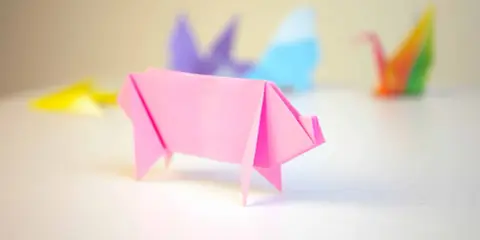 Cerdo de origami rosa con otras figuras de papiroflexia desenfocadas al fondo.