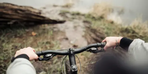 Ręce na kierownicy roweru, błotnista ścieżka i widok na wodę. To właśnie dlatego warto jeździć rowerem – bliskość natury i wolność.