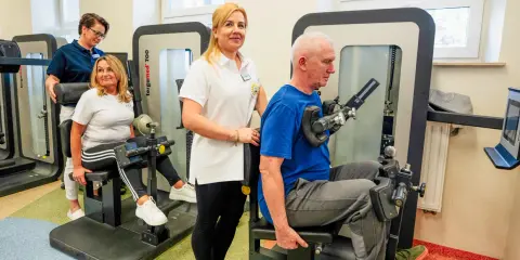 Rehabilitacja w Busko Zdrój: pan ćwiczy na siłowni pod okiem fizjoterapeutki, a pani z pomocą drugiej terapeutki. Sprawdź nasze sanatoria Busko Zdrój promocje!