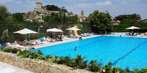 Piscina azzurra in un villaggio famiglie toscana, con castello medievale sullo sfondo e persone che si rilassano al sole.