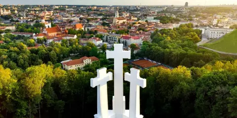 Wzgórze Trzech Krzyży w Wilnie z widokiem na miasto. Trzy białe krzyże górują nad zielonymi drzewami i zabytkową architekturą.