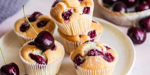 Saftige Muffins mit Sauerkirschen, bestäubt mit Puderzucker. Frische Kirschen liegen daneben und auf einem Muffin.
