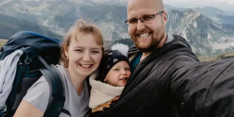 Rodzina na tle górskich szczytów. Uśmiechnięci rodzice z dzieckiem w nosidełku podziwiają czerwone wierchy.