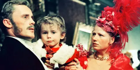 Rodzina w bogatych strojach, ojciec trzyma dziecko. Scena z filmu "Ziemia obiecana" streszczenie.