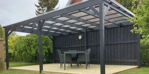 En mörk paviljong på en träaltan med bord och stolar. Perfekt för fika i trädgården.