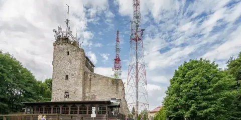 Kamienna wieża i wysokie maszty telekomunikacyjne na szczycie Chełmiec. Ludzie odpoczywają na trawie.