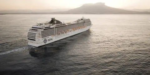 Kryssningsfartyget MSC Poesia glider fram på havet i solnedgången, med en vulkan i bakgrunden.