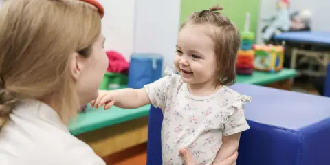 Uśmiechnięte dziecko w białej bluzce z kwiatkami wyciąga rączkę do terapeuty.