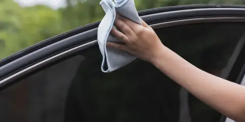 Mano limpiando los cristales del coche con un paño de microfibra azul. Un paso esencial para saber cómo limpiar los cristales del coche y mantener una visibilidad perfecta.