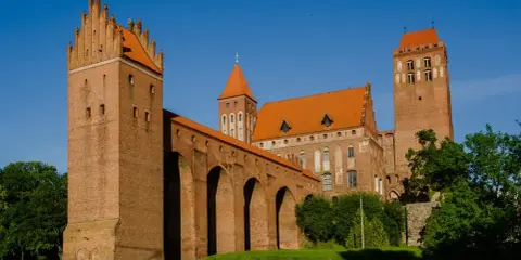 Potężny zamek w Kwidzynie z cegły, z charakterystycznymi wieżami i arkadowym murem obronnym, lśni w słońcu na tle błękitnego nieba.