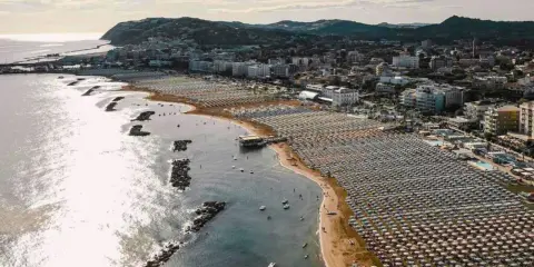 Vista aerea della cattolica spiaggia, con file ordinate di ombrelloni e lettini che si estendono lungo la costa, mentre il mare luccica sotto il sole.