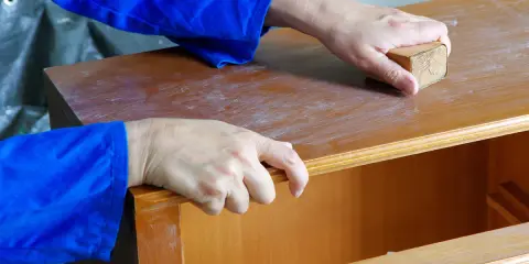 Hände schleifen eine Holzoberfläche mit einem Schleifklotz. So lernt man, wie lackiere ich richtig für ein glattes Ergebnis.