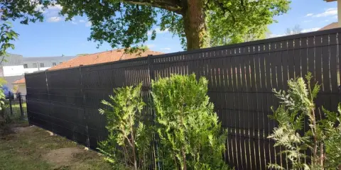 Clôture ajourée noire, offrant une protection discrète pour les tuyaux PVC. Des arbustes verts bordent cette barrière moderne.