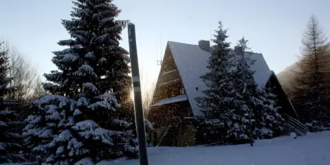 Zimowy krajobraz z bacówką pod Małą Rawką. Śnieg sypie, a słońce przebija się przez drzewa.
