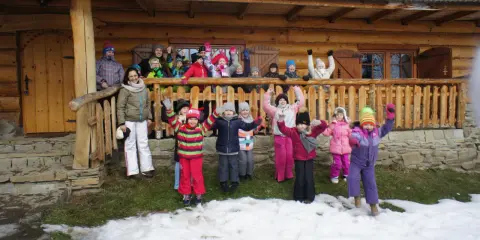 Wesoła gromadka dzieci pozdrawia z bacówki u harnasia. Wszyscy ubrani w zimowe stroje, radośnie machają rękami na tle drewnianego domu.
