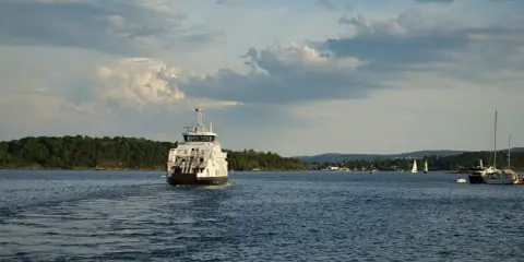 Un ferry blanc navigue sur une eau bleue, laissant une traînée derrière lui. La vitesse du bateau est visible dans le sillage. Au loin, des voiliers et une côte boisée.