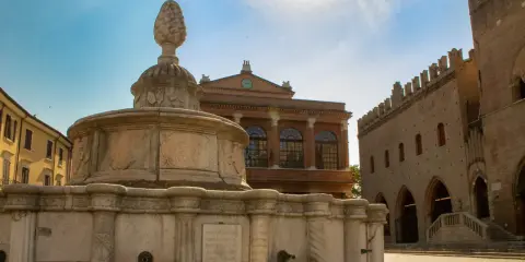 Fontana in pietra con pigna, edifici storici e cielo azzurro. Un assaggio dei monumenti di Rimini.