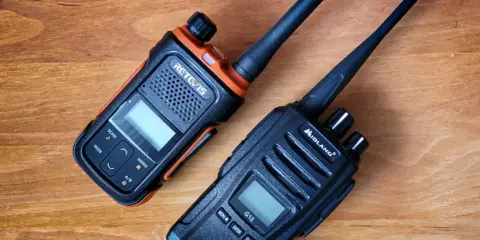 Dwa radiotelefony, jeden pomarańczowo-czarny RETC/IS, drugi czarny Midland G13, leżą na drewnianym blacie.