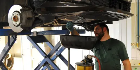 Mechanik w warsztacie wymienia zużyty olej samochodowy. Podniesiony pojazd i naczynie na płyn wskazują na rutynową czynność serwisową.
