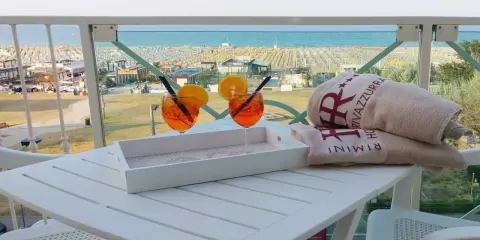 Due aperitivi arancioni e asciugamani con logo "Hotel Rivazzurra Rimini" sul tavolo, con vista sul rivazzurra mare e spiaggia attrezzata.
