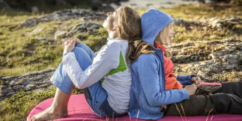 Två personer, en vuxen och ett barn, sitter rygg mot rygg på en liggunderlag i naturen. En härlig stund att vandra med barn.