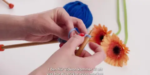 Händer stickar med garn och stickor. En blå garnboll och orange gerberor i bakgrunden. Lär dig stockinette stitch svenska.