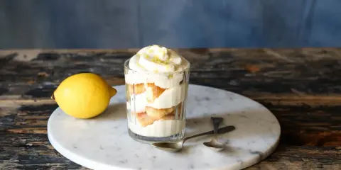 Ein Glas mit geschichtetem Zitronen Tiramisu, garniert mit Zitronenschale. Daneben liegt eine Zitrone.