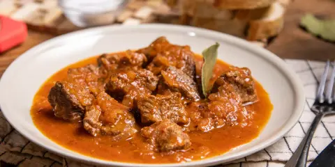 Sabrosa carne con tomate, guisada lentamente hasta quedar tierna, servida en un plato blanco con pan y sal al lado.