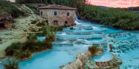 Cascate termali turchesi in Toscana, con un antico mulino in pietra e un cielo rosa al tramonto. Curiosità sulla Toscana: un luogo magico.