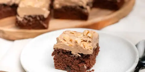 En bit chokladtårta med krämig frosting och nötter. Flera bitar serveras på ett träfat i bakgrunden.