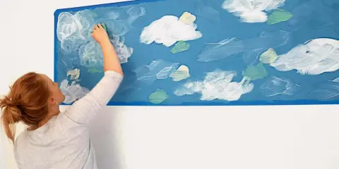 Kobieta maluje gąbką białe chmury na niebieskiej ścianie. Prosty sposób, jak namalować chmury na ścianie, tworząc niebo.