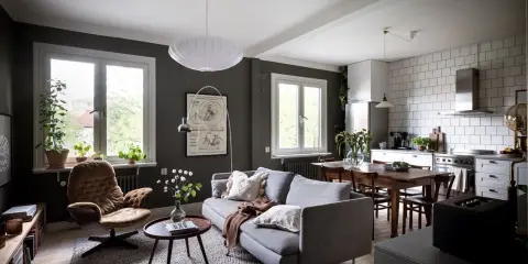 Un salón rectangular amueblado con sofá gris, sillón marrón, mesa de centro y alfombra. Cocina abierta con mesa de comedor.