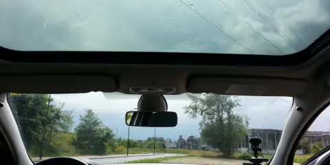 Widok z wnętrza samochodu z panoramicznym dachem, przez który widać niebo i drzewa. Krople deszczu na szybie.