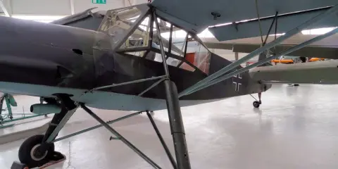 Ein altes Flugzeug, ein Storch, steht in einer Halle. Das Flugzeug hat eine Tarnlackierung und ein Kreuz auf dem Heck.