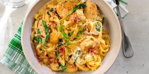 En skål med krämig pasta med soltorkade tomater, kyckling och spenat, toppad med riven parmesan och färska örter.