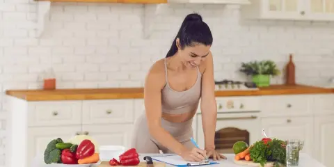 Mujer sonriente planifica su dieta vegetariana para deportistas en la cocina, rodeada de verduras frescas.