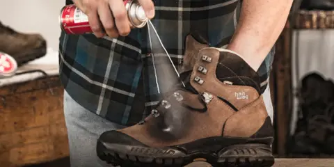 Jak zabezpieczyć nowe buty skórzane? Mężczyzna spryskuje brązowe buty trekkingowe preparatem ochronnym.