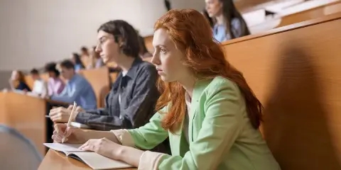 Studentka z rudymi włosami skupiona na wykładzie, robi notatki. Jej kolega obok również studiuje psychologię.