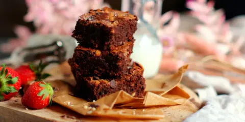 Saftige Brownies nach original Rezept, gestapelt auf einem Holzbrett mit Erdbeeren und einem Krug Milch.