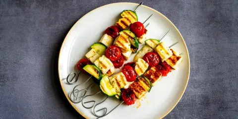 Brochettes de barbecue végétarien : cubes de halloumi grillé, tomates cerises et courgettes sur des brochettes métalliques, servis sur une assiette.
