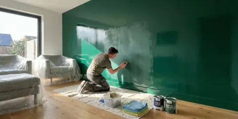 Hombre lijando pared verde brillante, preparándose para pintar. Muebles cubiertos, latas de pintura a mano.