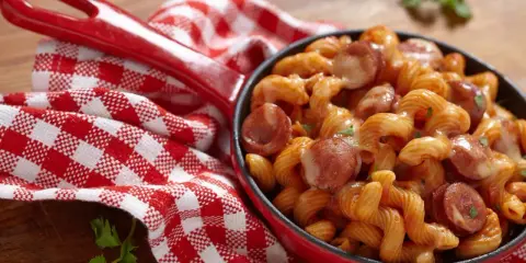 Pyszne spaghetti z kiełbasą w sosie pomidorowym, podane w czerwonej patelni, obok czerwono-białej kratkowanej serwetki.